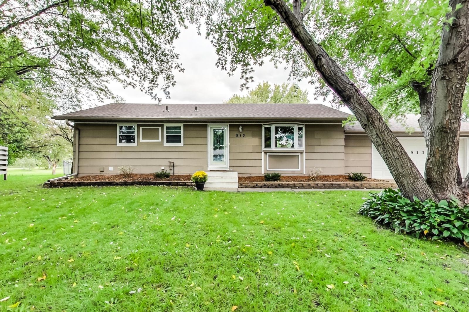 Front of a single-storey tan house with a bay window, wide front lawn and mature shade trees.
