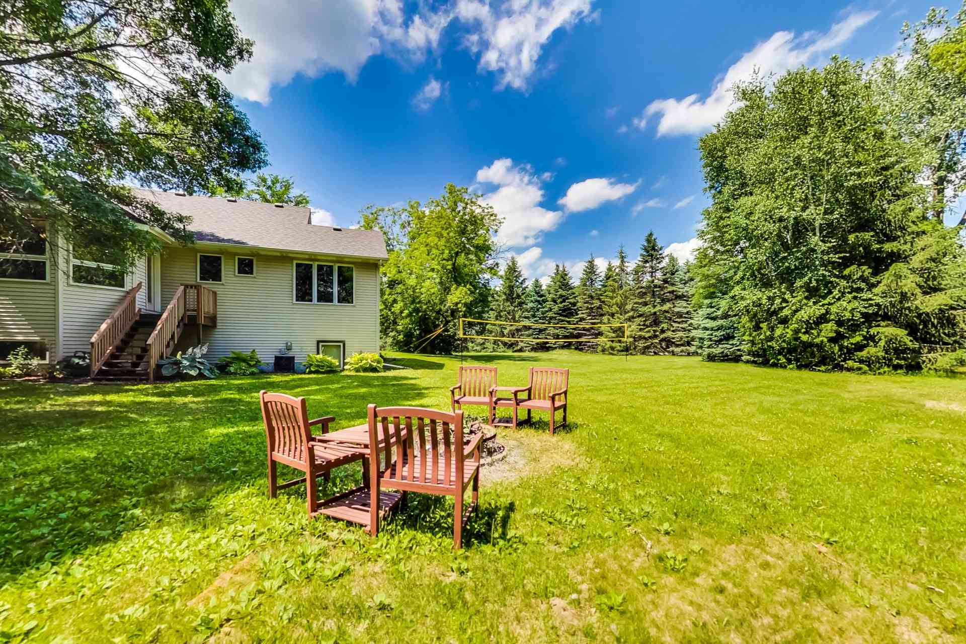 Back yard with a wide mown lawn, a volleyball net, fire-pit chairs and stairs up to the deck.